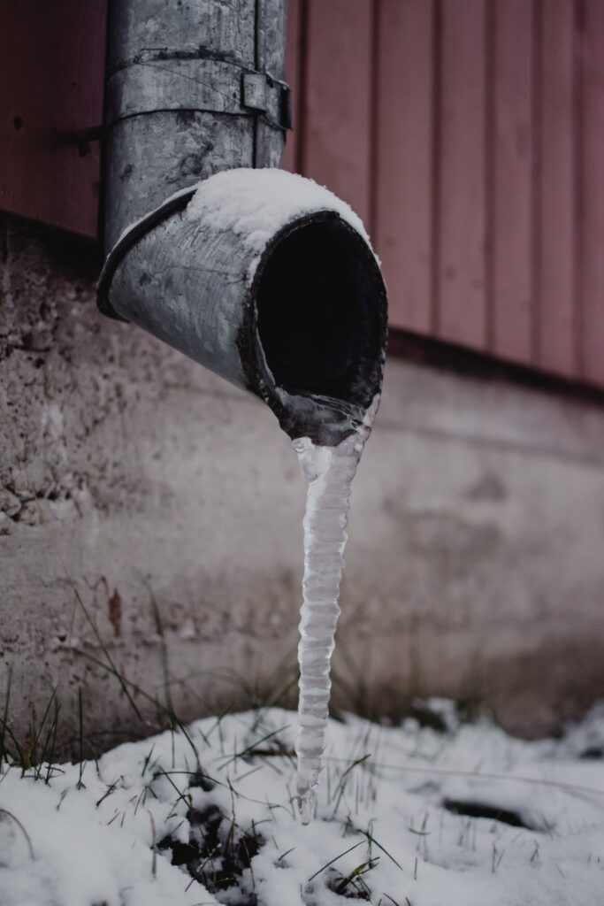 Icicle hanging from a metal downspout in a snowy yard, illustrating potential plumbing issues related to drainage in winter conditions.