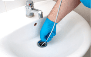 Person in blue gloves using a plumbing snake to clear a clogged sink drain, highlighting plumbing repair services offered by Frost & Kretsch Plumbing Inc. in Roseville.