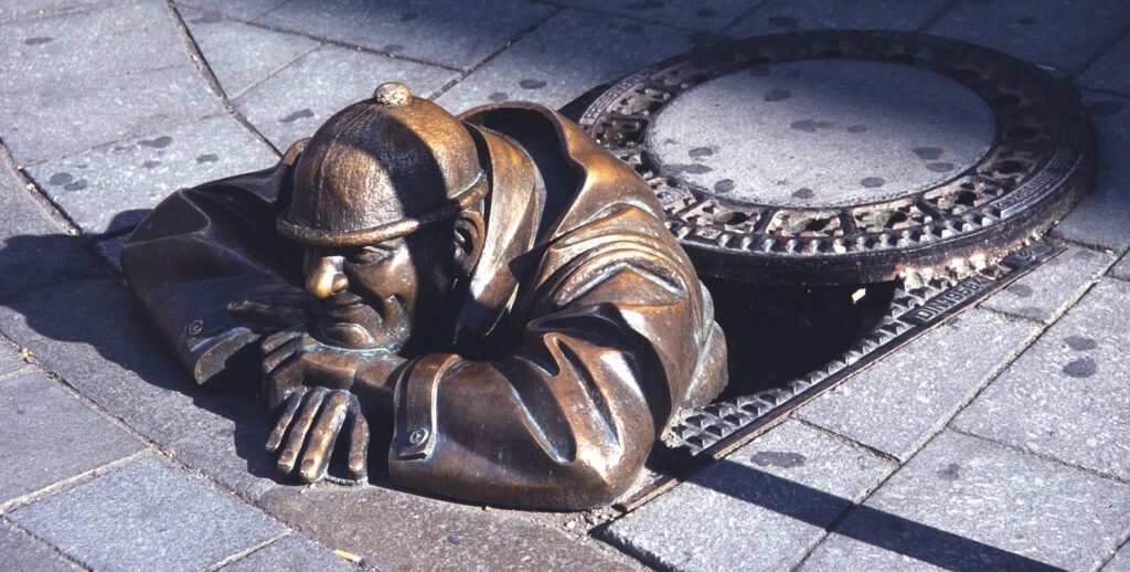 Bronze sculpture of a man in a hat peering out from a manhole cover on a city street, symbolizing plumbing work and local expertise in Utica.