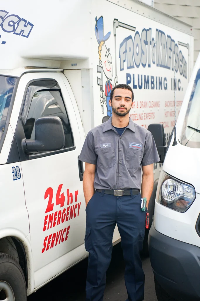 Plumber standing between two service vans, one featuring "24 hr Emergency Service.